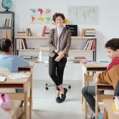 Nauczycielka w eleganckim stroju, zgodnym z dress code'em nauczyciela, stoi przed klasą. Uczniowie siedzą w ławkach.
