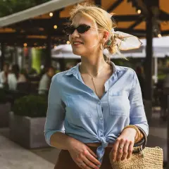 Kobieta w błękitnej, zawiązanej na supeł bluzce, okularach i z torebką ze słomy.