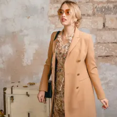 Elegancka kobieta w płaszczu camelowy kolor, sukience w kwiaty i okularach przeciwsłonecznych stoi obok starej skrzyni.