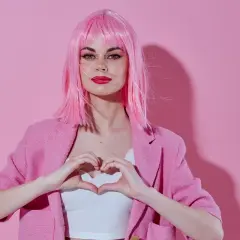 Kobieta w różowej peruce i marynarce, tworząca serce dłońmi. Całość w stylu barbiecore.