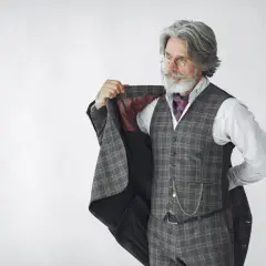 Starszy pan z siwą brodą i wąsami, ubrany w kraciasty garnitur, kamizelkę i koszulę, prezentuje się jako arbiter elegancji.