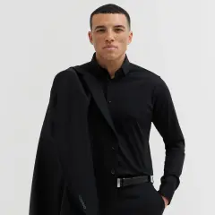 Mężczyzna w czarnym garniturze i koszuli, z marynarką na ramieniu. Elegancki, czarny pasek do garnituru podkreśla jego styl.