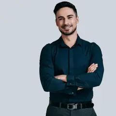 Elegancki mężczyzna w granatowej koszuli i szarych spodniach, z rękami skrzyżowanymi na piersi. Jego stylizacje są zawsze nienaganne.