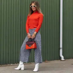 Stylowa kobieta w pomarańczowym golfie i spodniach culotte w kratkę, białych kozakach i okularach.