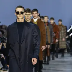 Model w ciemnym płaszczu i okularach na wybiegu, w tle inni modele i publiczność.