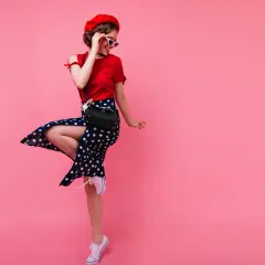 Dziewczyna w stylu pin-up girl w czerwonej bluzce, spódnicy w grochy i czerwonym beretcie skacze na różowym tle.