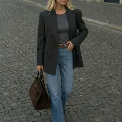 Elegancka kobieta w szarym longsleeve, jeansach i marynarce idzie po bruku, niosąc brązową torebkę.