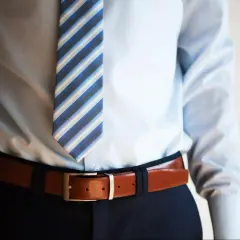 Elegancki mężczyzna w jasnej koszuli i granatowych spodniach pokazuje, jak nosić pasek męski. Brązowy pasek z prostą klamrą idealnie podkreśla talię.