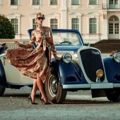 Elegancka kobieta w kwiecistej sukience i okularach przeciwsłonecznych opiera się o zabytkowy samochód. Stylowe lata 20. w pełnej krasie.