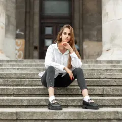 Kobieta w białej koszuli i szarych jeansach siedzi na schodach, prezentując czarne mokasyny.