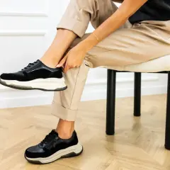 Kobieta w beżowych spodniach i czarnych sneakersach na grubej podeszwie siedzi na krześle.