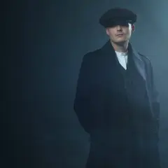 Mężczyzna w kaszkiecie i płaszczu, styl Peaky Blinders, z rękami w kieszeniach, w zadymionym otoczeniu.