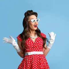 Uśmiechnięta pin-up girl w czerwonej sukience w grochy, białych rękawiczkach i okularach, z uniesionymi rękami.
