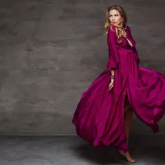 Kobieta w zwiewnej sukni w kolorze amarantowym wiruje na tle szarej ściany.