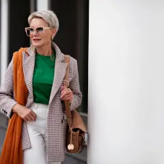 Elegancka kobieta po 60. w stylowej marynarce, zielonej bluzce i białych spodniach. Pomarańczowy szal dodaje jej uroku.