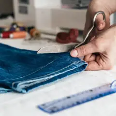 Ręce tną dżinsy nożyczkami, przygotowując je do skrócenia. Obok leży linijka i szpulki nici.