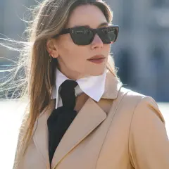Elegancka kobieta w beżowym płaszczu, okularach przeciwsłonecznych i białej koszuli z czarną apaszką, tworzącą ciekawy rodzaj kołnierzyka.