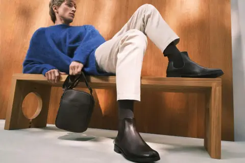 Eleganckie, ciemne botki to idealne buty do chinosów i koszuli. Mężczyzna w niebieskim swetrze prezentuje stylowy zestaw.