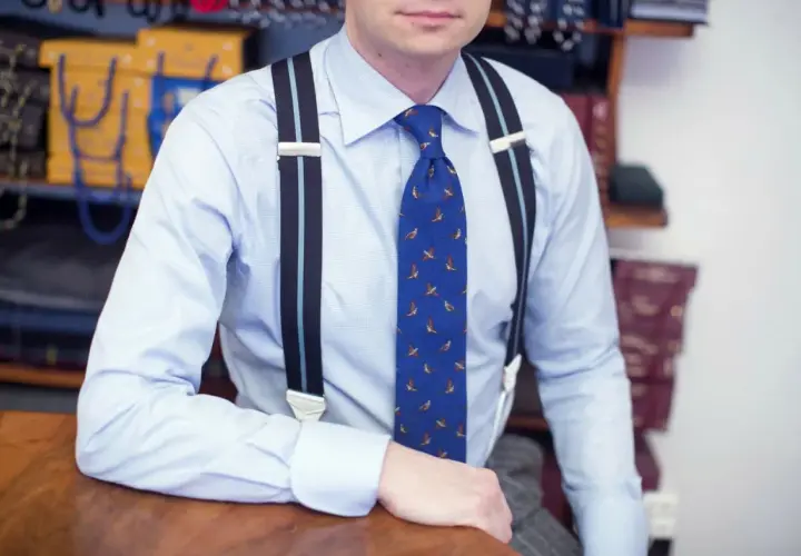 Elegancki mężczyzna w błękitnej koszuli i krawacie z ptakami, pokazuje, jak nosić szelki.