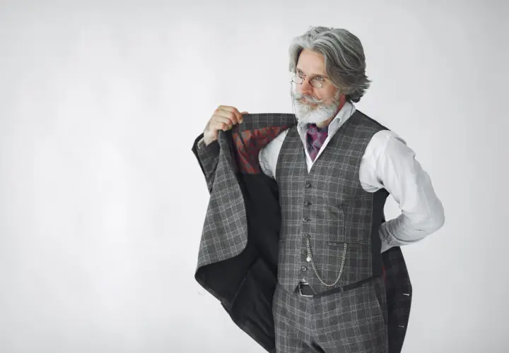 Starszy pan z siwą brodą i wąsami, ubrany w kraciasty garnitur, kamizelkę i koszulę, prezentuje się jako arbiter elegancji.