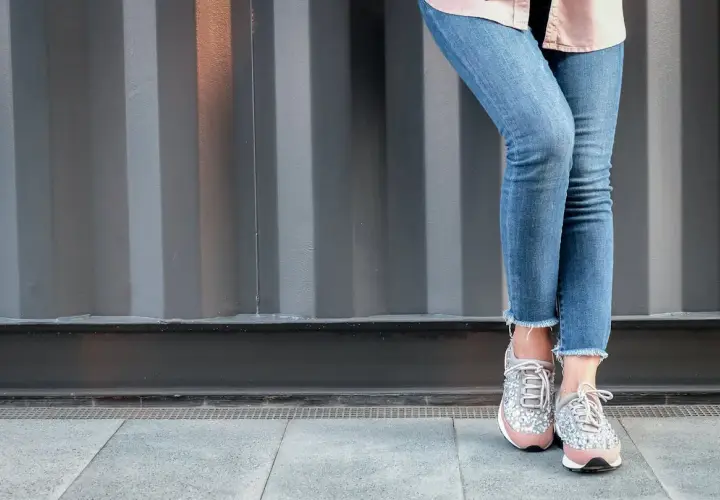 Nogawki jeansów postrzępione na dole, modne sneakersy z błyszczącymi detalami.