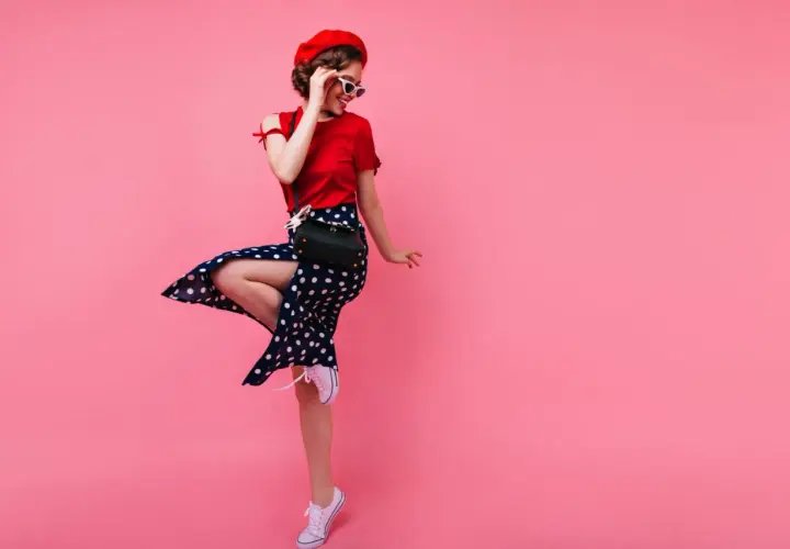 Dziewczyna w stylu pin-up girl w czerwonej bluzce, spódnicy w grochy i czerwonym beretcie skacze na różowym tle.