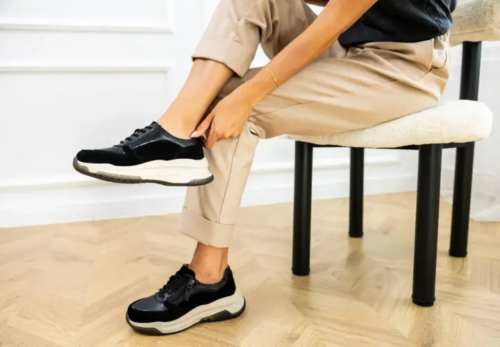Kobieta w beżowych spodniach i czarnych sneakersach na grubej podeszwie siedzi na krześle.