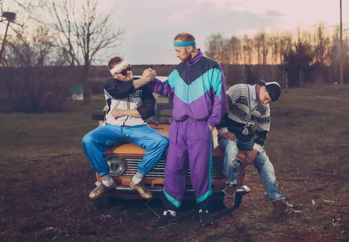 Trzech mężczyzn w dresach i okularach przeciwsłonecznych siedzi na masce starego samochodu. Stylizacja rodem z lat 90. xx wieku.