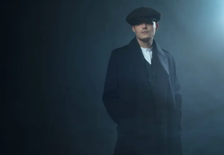 Mężczyzna w kaszkiecie i płaszczu, styl Peaky Blinders, z rękami w kieszeniach, w zadymionym otoczeniu.