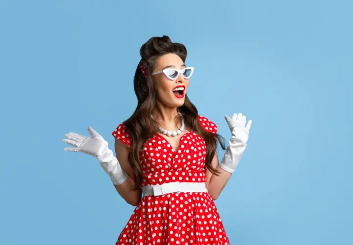 Uśmiechnięta pin-up girl w czerwonej sukience w grochy, białych rękawiczkach i okularach, z uniesionymi rękami.