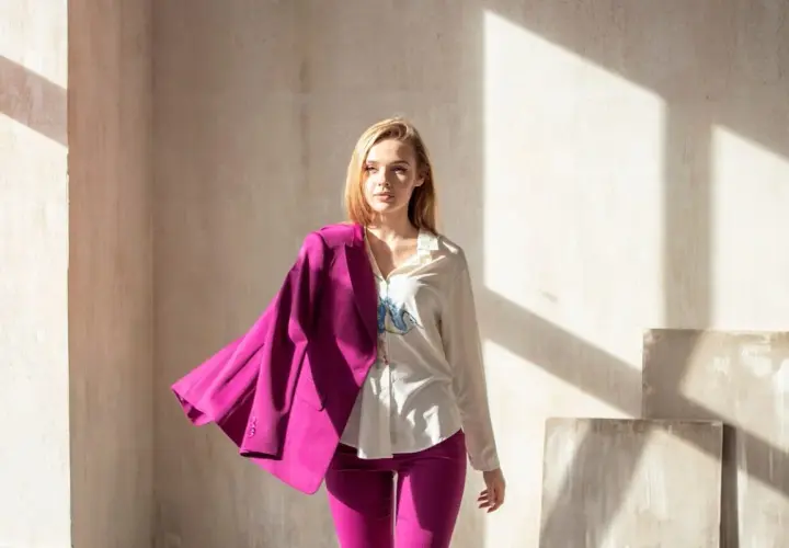 Kobieta w stylizacji viva magenta, z marynarką narzuconą na ramię, stoi w świetle słonecznym.