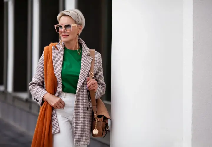 Elegancka kobieta po 60. w stylowej marynarce, zielonej bluzce i białych spodniach. Pomarańczowy szal dodaje jej uroku.