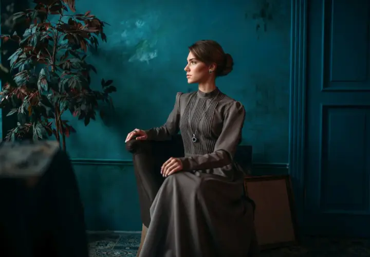 Elegancka kobieta w sukni w stylu light academia siedzi w fotelu, w tle ciemnozielona ściana i roślina.