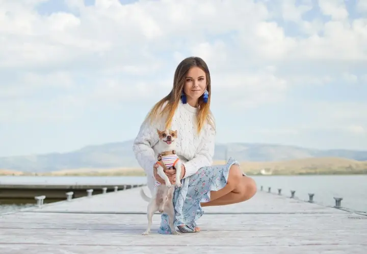 Dziewczyna w swetrze i kwiecistej sukience, z chihuahuą na rękach, prezentuje swój letni styl ubierania na tle jeziora.