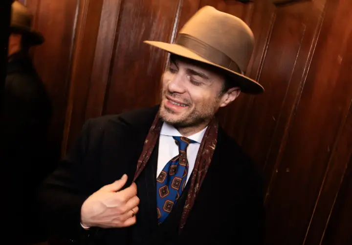 Elegancki mężczyzna w brązowym kapeluszu, czarnym płaszczu i stylowym krawacie. Pokazuje, jak nosić kapelusz męski, dodając klasy każdej stylizacji.