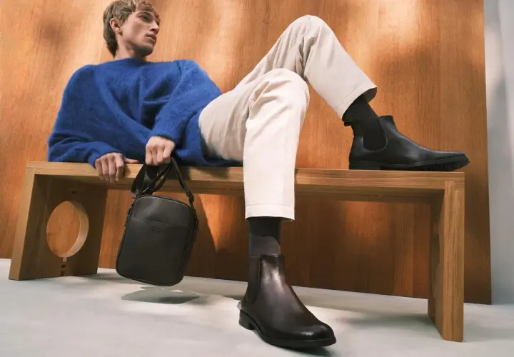 Eleganckie, ciemne botki to idealne buty do chinosów i koszuli. Mężczyzna w niebieskim swetrze prezentuje stylowy zestaw.