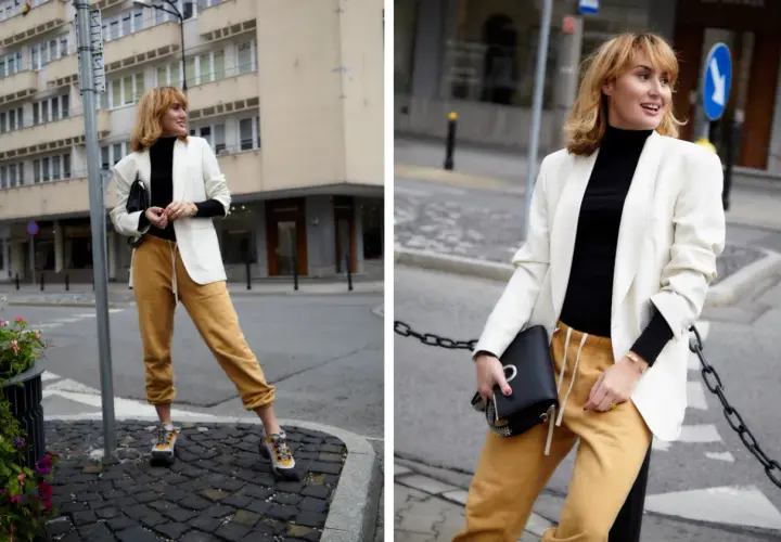 Modna kobieta w stylizacji z czarnym golfem, białej marynarce i musztardowych spodniach dresowych.