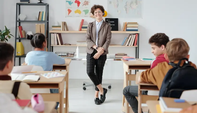 Nauczycielka w eleganckim stroju, zgodnym z dress code'em nauczyciela, stoi przed klasą. Uczniowie siedzą w ławkach.