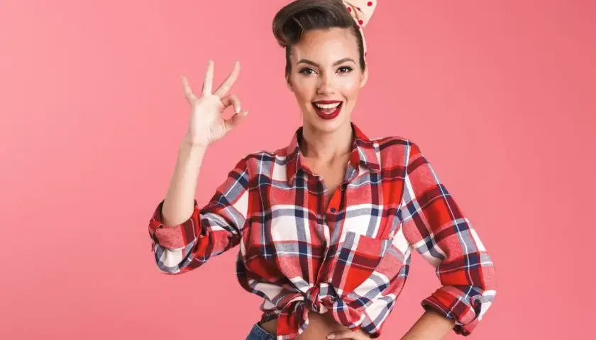 Uśmiechnięta kobieta w stylu pin-up, w kratowanej koszuli i dżinsach, pokazuje znak "OK".