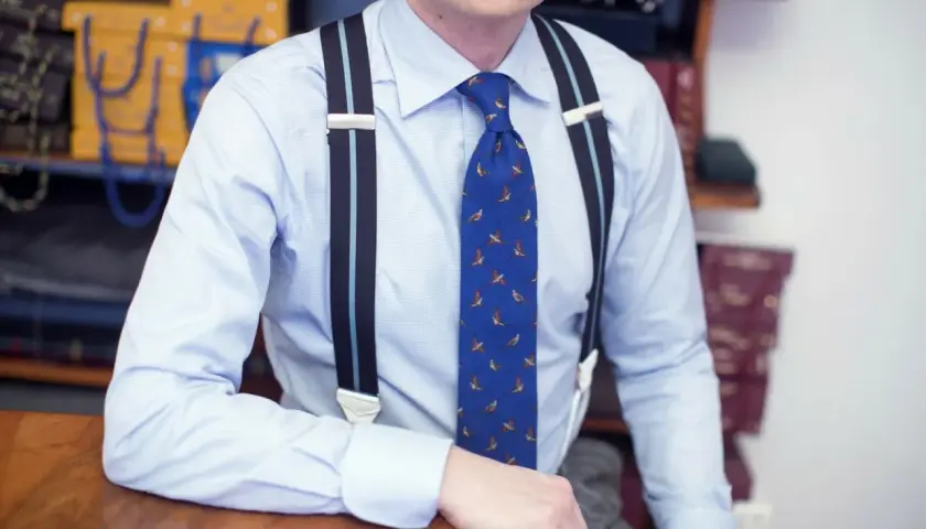 Elegancki mężczyzna w błękitnej koszuli i krawacie z ptakami, pokazuje, jak nosić szelki.
