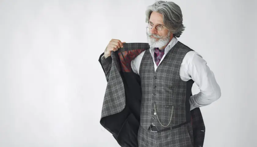 Starszy pan z siwą brodą i wąsami, ubrany w kraciasty garnitur, kamizelkę i koszulę, prezentuje się jako arbiter elegancji.