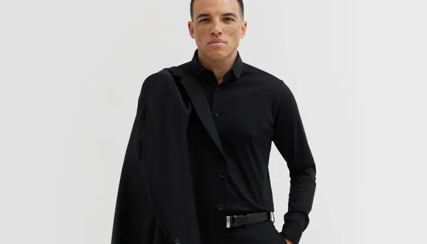 Mężczyzna w czarnym garniturze i koszuli, z marynarką na ramieniu. Elegancki, czarny pasek do garnituru podkreśla jego styl.