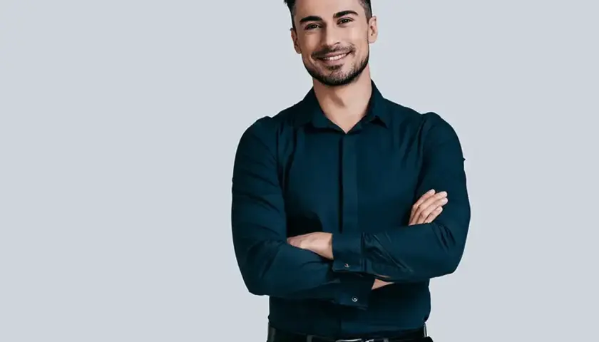Elegancki mężczyzna w granatowej koszuli i szarych spodniach, z rękami skrzyżowanymi na piersi. Jego stylizacje są zawsze nienaganne.