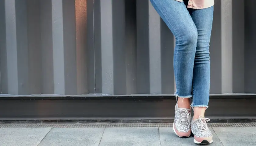 Nogawki jeansów postrzępione na dole, modne sneakersy z błyszczącymi detalami.