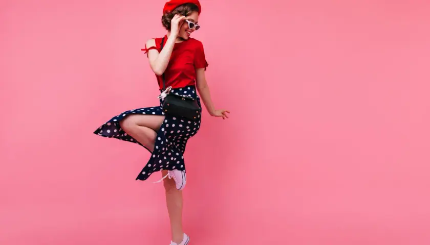 Dziewczyna w stylu pin-up girl w czerwonej bluzce, spódnicy w grochy i czerwonym beretcie skacze na różowym tle.