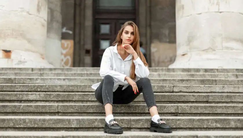 Kobieta w białej koszuli i szarych jeansach siedzi na schodach, prezentując czarne mokasyny.