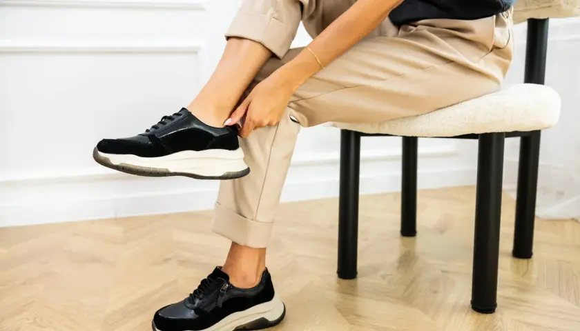 Kobieta w beżowych spodniach i czarnych sneakersach na grubej podeszwie siedzi na krześle.