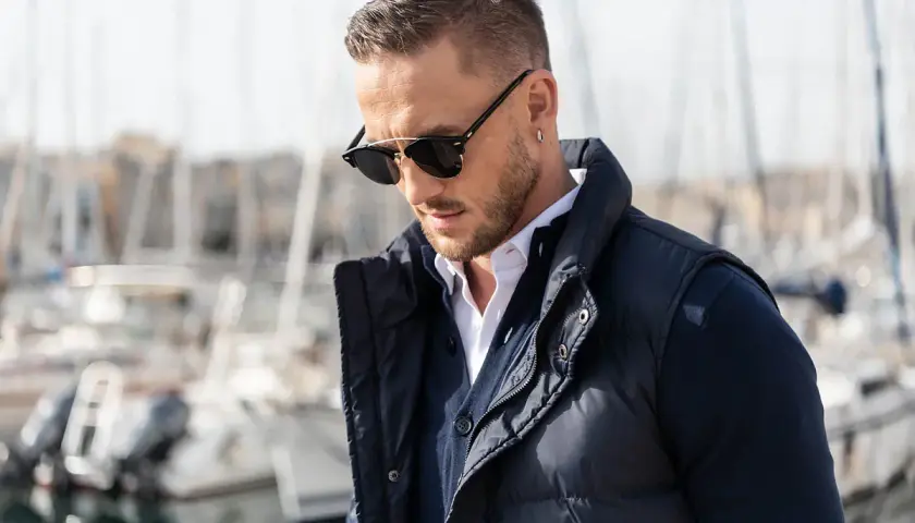Mężczyzna w okularach przeciwsłonecznych, białej koszuli i granatowej kamizelce, prezentujący styl country męski. W tle port z łodziami.