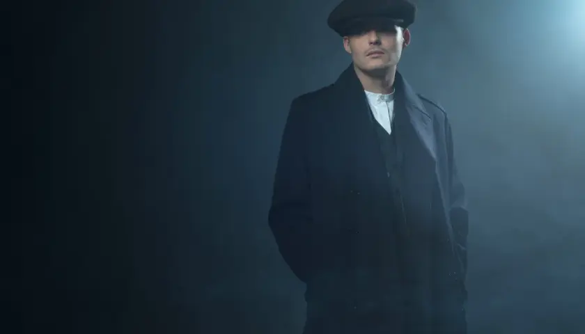 Mężczyzna w kaszkiecie i płaszczu, styl Peaky Blinders, z rękami w kieszeniach, w zadymionym otoczeniu.