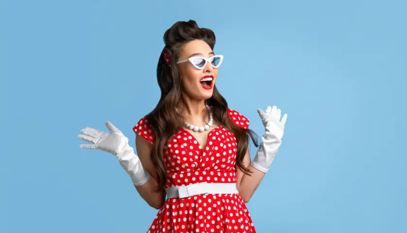 Uśmiechnięta pin-up girl w czerwonej sukience w grochy, białych rękawiczkach i okularach, z uniesionymi rękami.